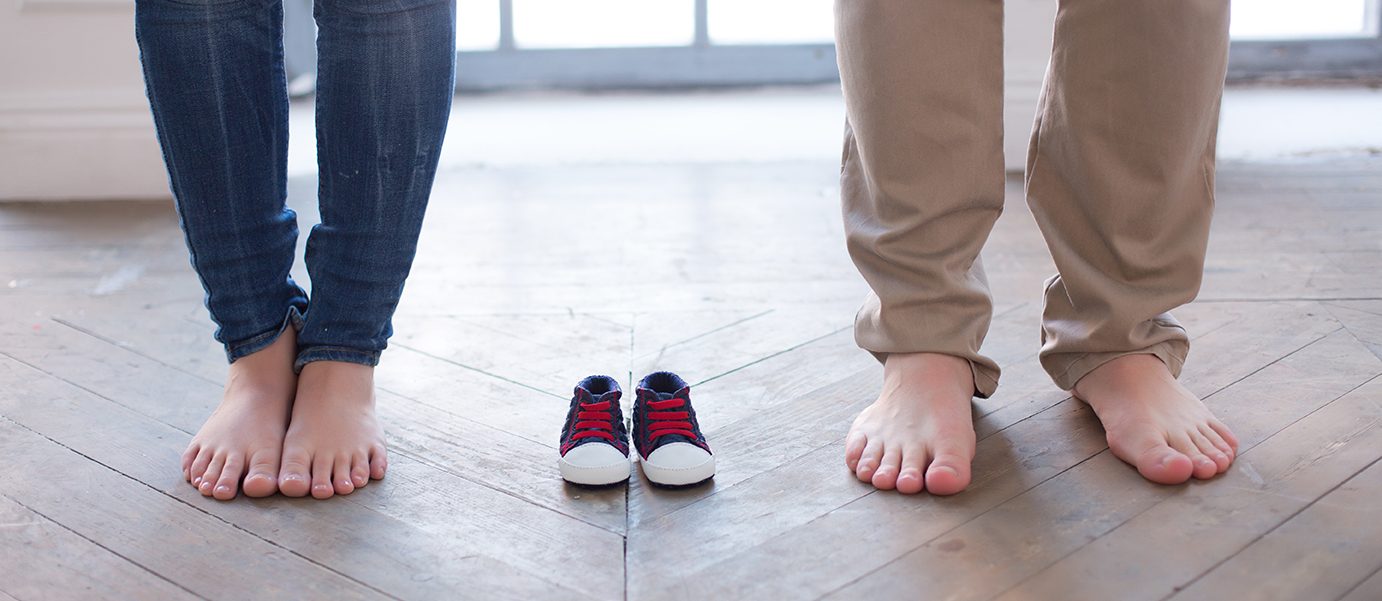 A couple that's expecting a baby stands with a pair of baby shoes between them